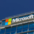 Microsoft corporate headquarters building with the Microsoft logo prominently displayed on a modern office exterior. The image features the iconic four-colored square logo (red, green, blue, and yellow) next to the company's name in bold white letters, set against a glass façade with blue-tinted windows.