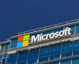 Microsoft corporate headquarters building with the Microsoft logo prominently displayed on a modern office exterior. The image features the iconic four-colored square logo (red, green, blue, and yellow) next to the company's name in bold white letters, set against a glass façade with blue-tinted windows.