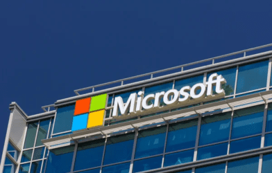 Microsoft corporate headquarters building with the Microsoft logo prominently displayed on a modern office exterior. The image features the iconic four-colored square logo (red, green, blue, and yellow) next to the company's name in bold white letters, set against a glass façade with blue-tinted windows.