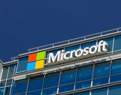 Microsoft corporate headquarters building with the Microsoft logo prominently displayed on a modern office exterior. The image features the iconic four-colored square logo (red, green, blue, and yellow) next to the company's name in bold white letters, set against a glass façade with blue-tinted windows.