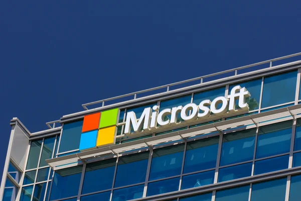 Microsoft corporate headquarters building with the Microsoft logo prominently displayed on a modern office exterior. The image features the iconic four-colored square logo (red, green, blue, and yellow) next to the company's name in bold white letters, set against a glass façade with blue-tinted windows.