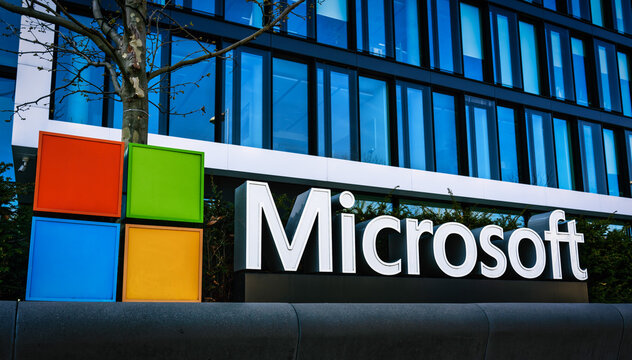 Microsoft’s 2025 Layoff - Microsoft corporate headquarters building with the Microsoft logo prominently displayed on a modern office exterior. The image features the iconic four-colored square logo (red, green, blue, and yellow) next to the company's name in bold white letters, set against a glass façade with blue-tinted windows.